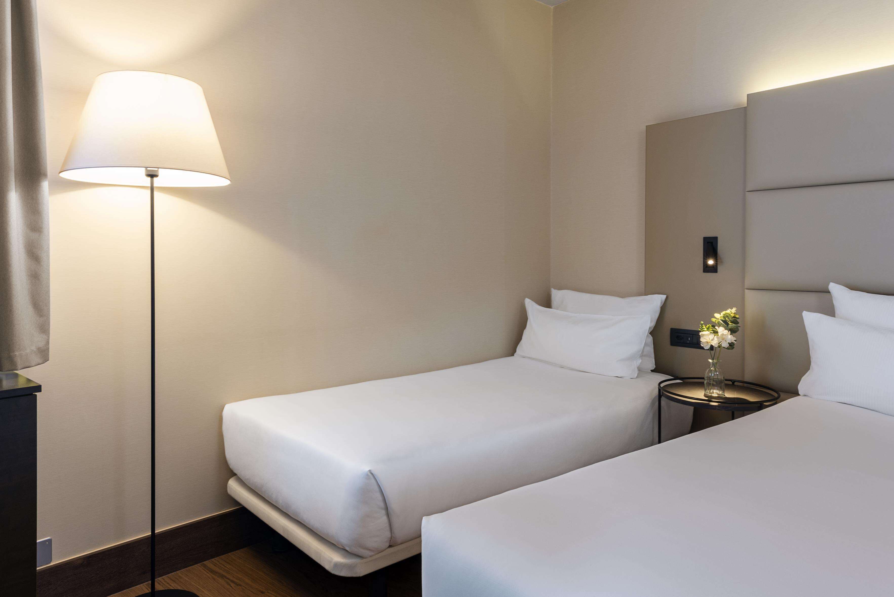 A hotel room with two single beds, a bedside table with a vase of flowers, a floor lamp, and a large mirror on the wall.