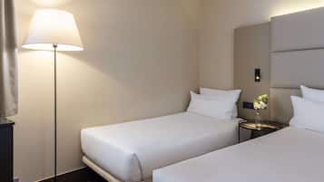 A hotel room with two single beds, a bedside table with a vase of flowers, a floor lamp, and a large mirror on the wall.