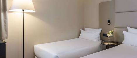 A hotel room with two single beds, a bedside table with a vase of flowers, a floor lamp, and a large mirror on the wall.