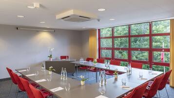 A conference room with a long table, red chairs, and large windows with a view of greenery.