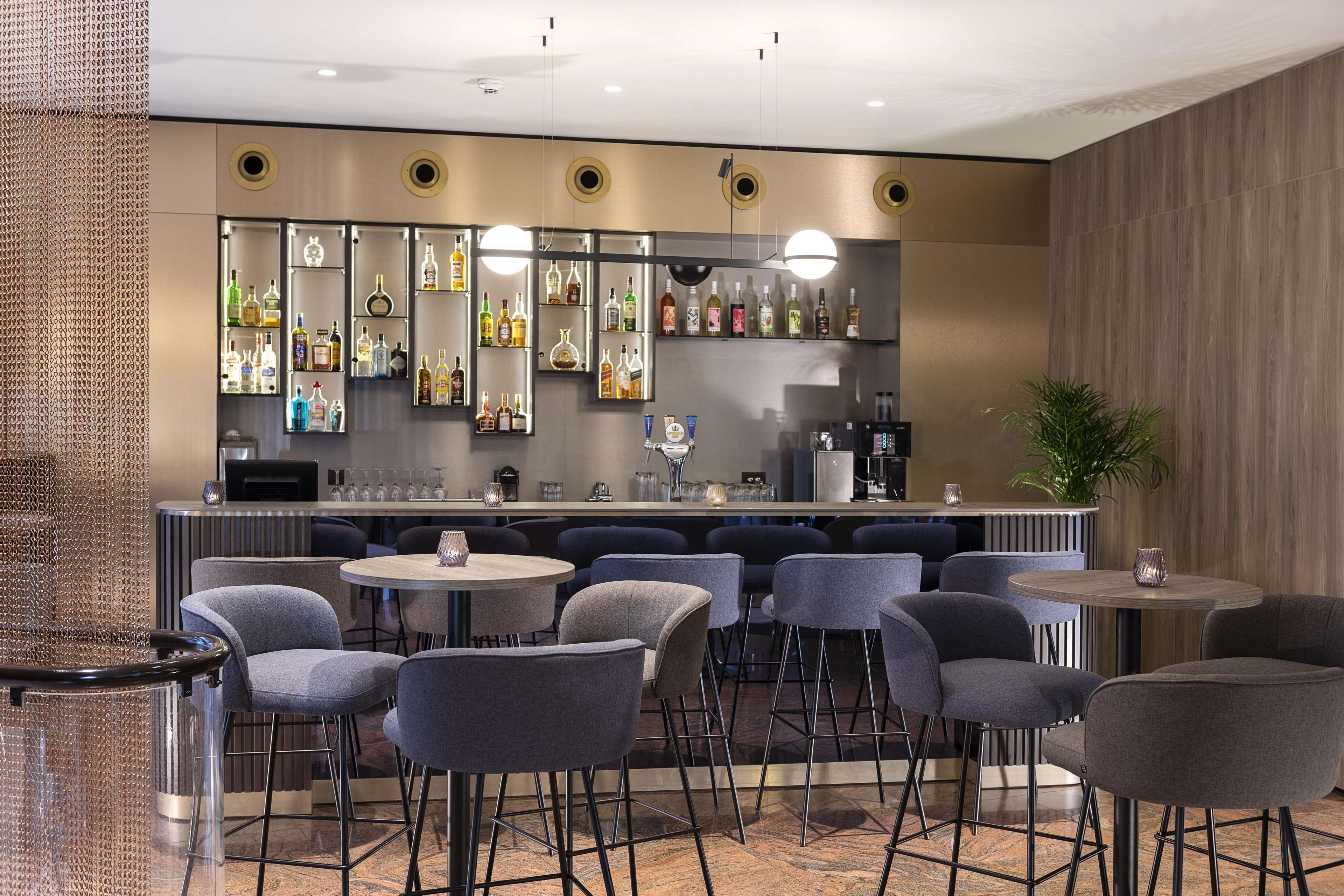 A modern bar area with multiple seating options, including bar stools and round tables, and a variety of bottles displayed on shelves.