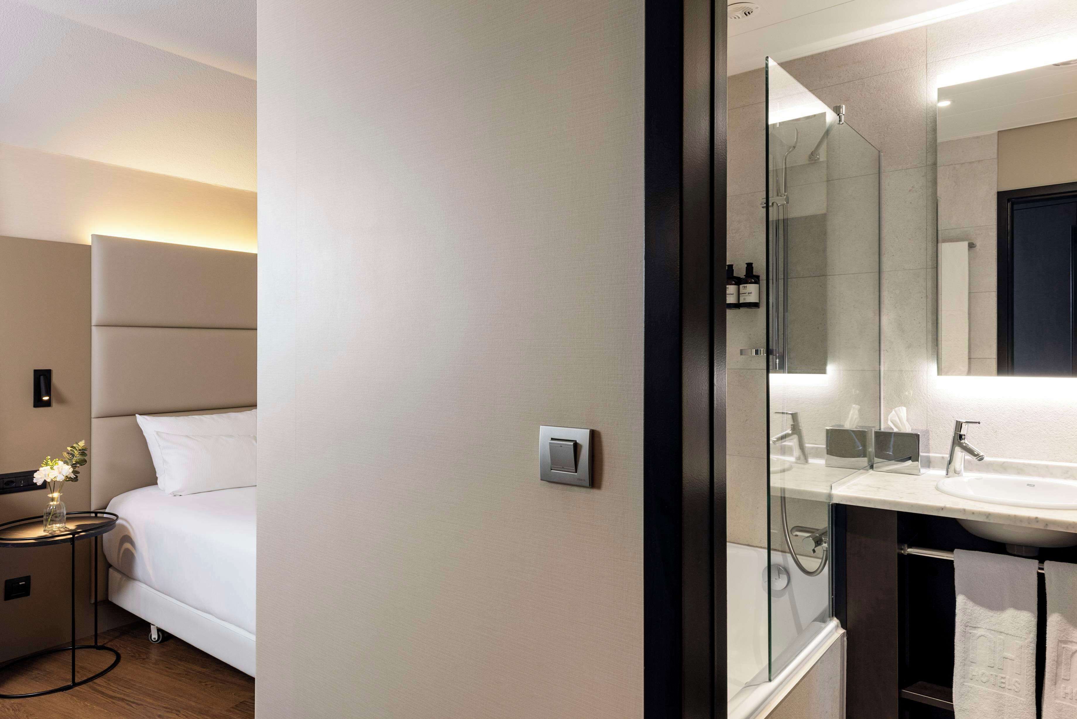 A modern hotel room with a bed, bedside table with a vase, a wall-mounted light switch, a glass shower enclosure, and a bathroom with a sink and towel rack.