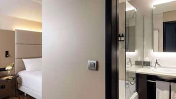 Una habitación de hotel moderna con cama, mesita de noche con un florero, interruptor de luz en la pared, cabina de ducha de vidrio y baño con lavamanos y toallero.