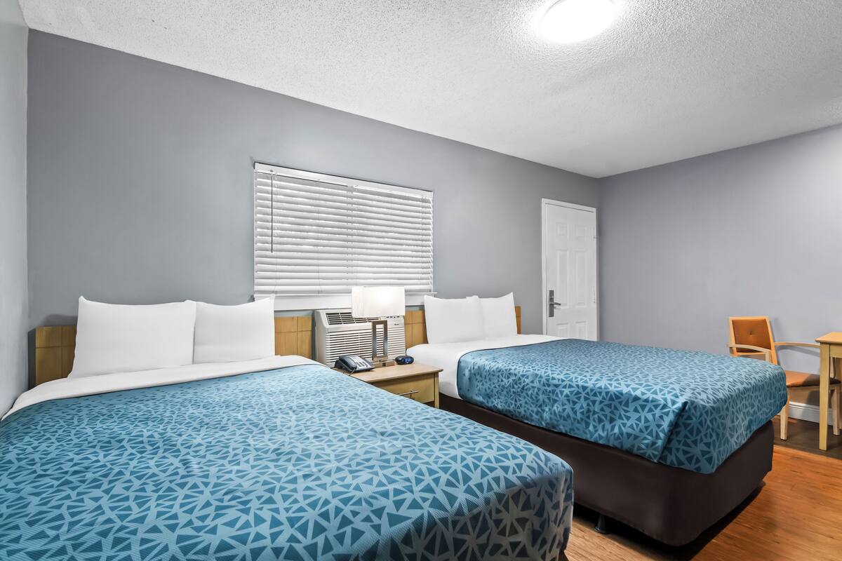 A hotel room with two beds, a desk, a chair, a window with blinds, and a door.