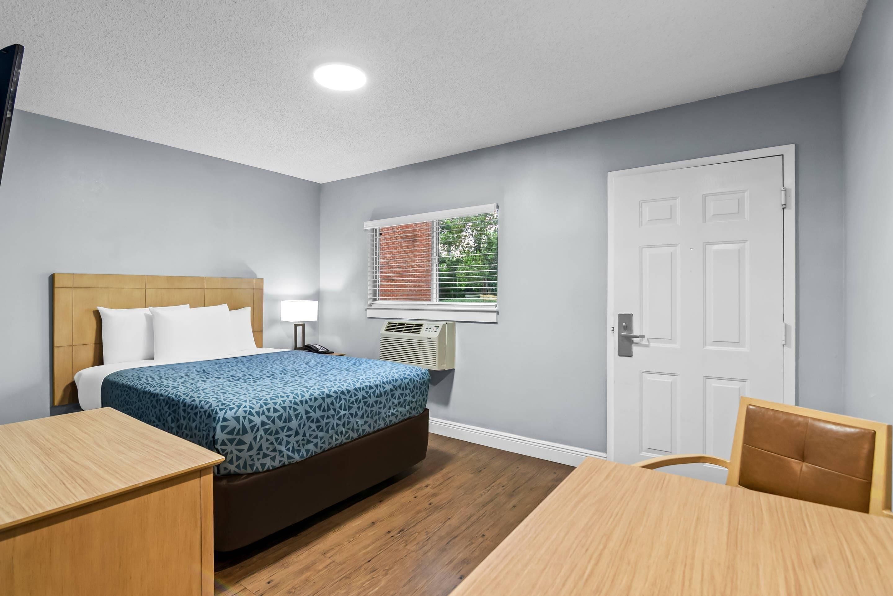 A hotel room with a bed, bedside table, chair, and a window with blinds.