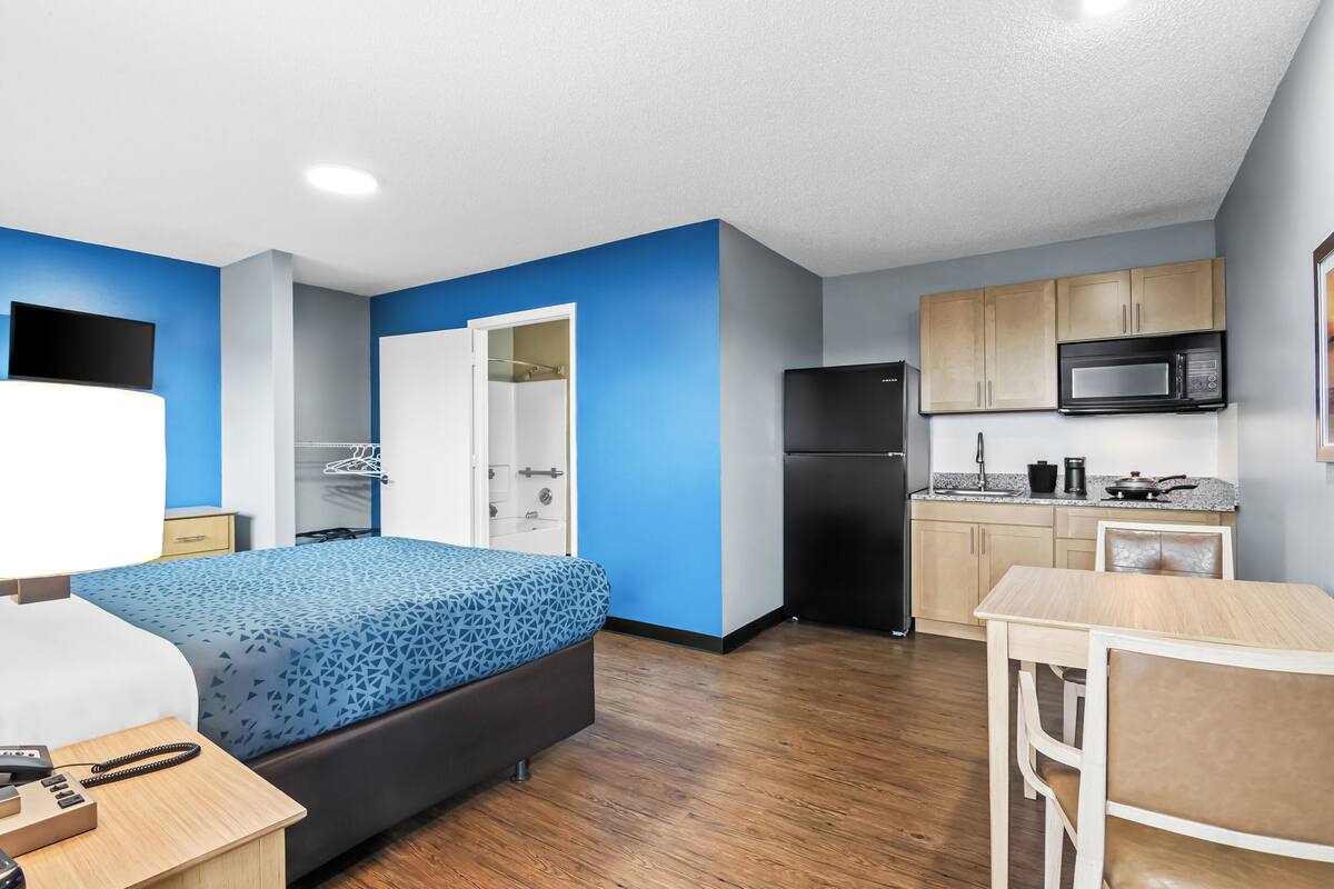 A compact hotel room with a bed, bedside table, and a small desk. A kitchenette with a microwave, stove, and cabinets is visible.