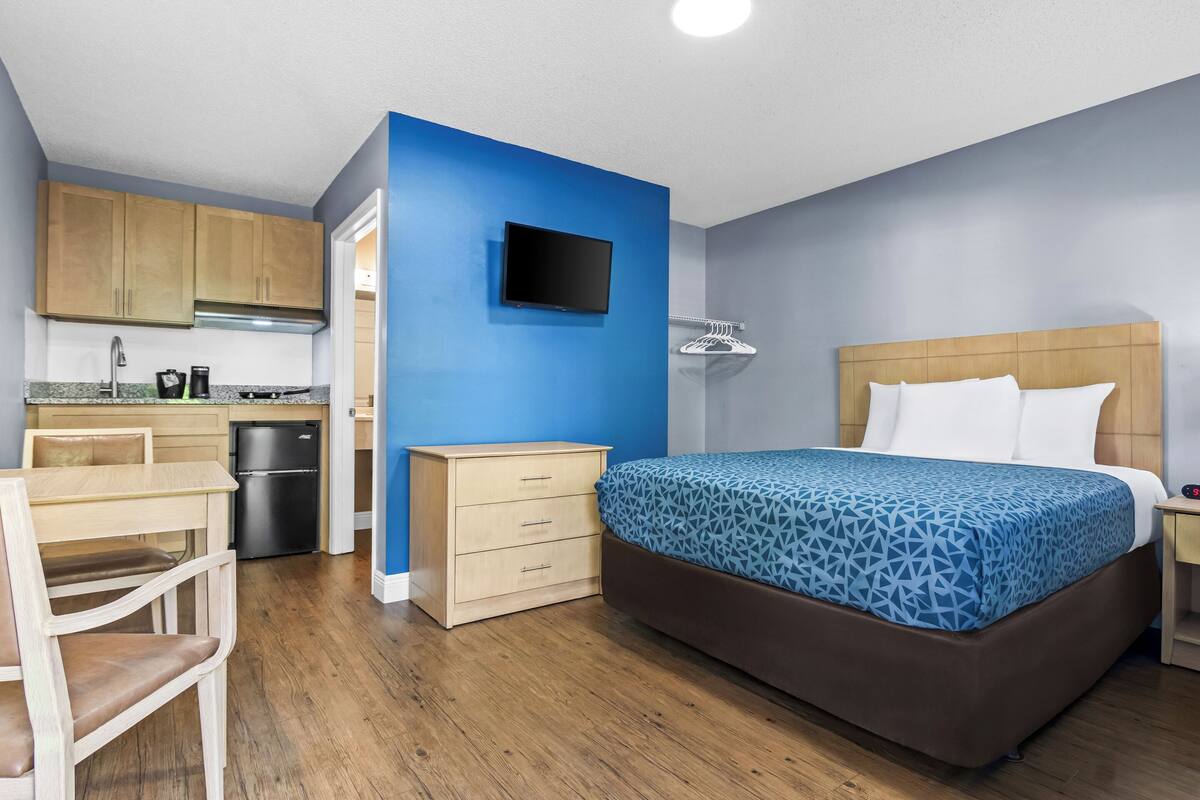 A compact hotel room with a bed, a small desk, a chair, a kitchenette, and a television.