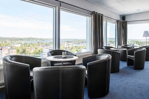 Habitación de hotel con un ventanal amplio que ofrece vistas a un lago y edificios. Hay varias sillas de cuero negro y una mesita redonda.