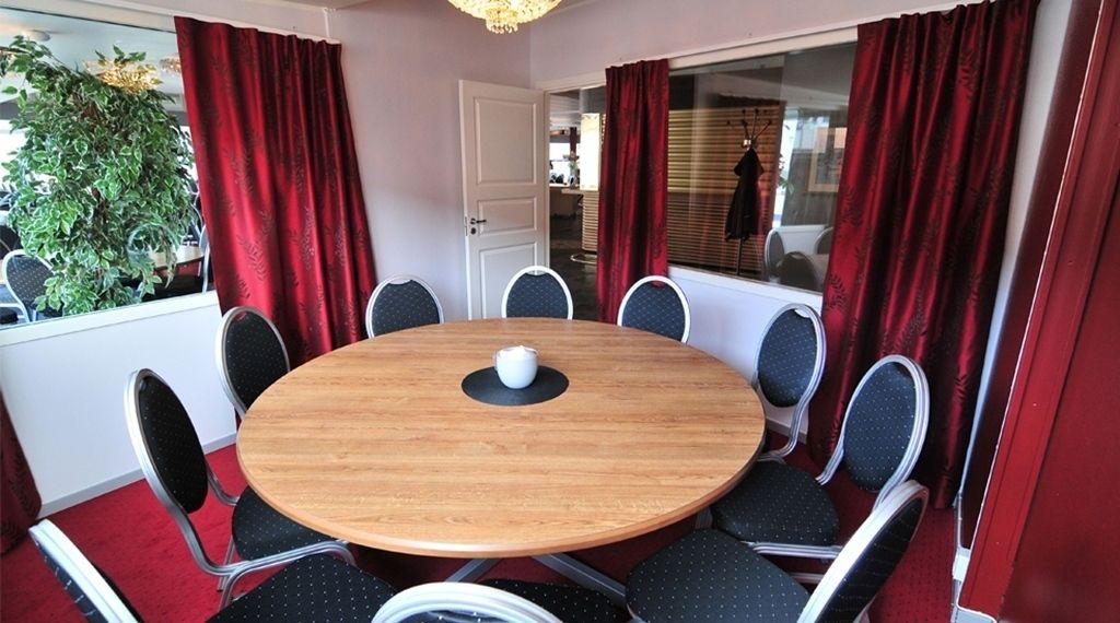 A round wooden table with eight chairs, red curtains, and a potted plant in the background.