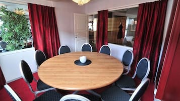 A round wooden table with eight chairs, red curtains, and a potted plant in the background.