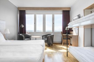 Habitación de hotel con dos camas, un escritorio, una silla, una lámpara y vistas a las montañas a través de amplias ventanas.