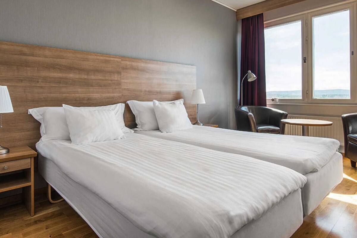 A hotel room with two beds, a wooden headboard, a desk with a lamp, a chair, a round table, and a view of the outdoors through a large window.