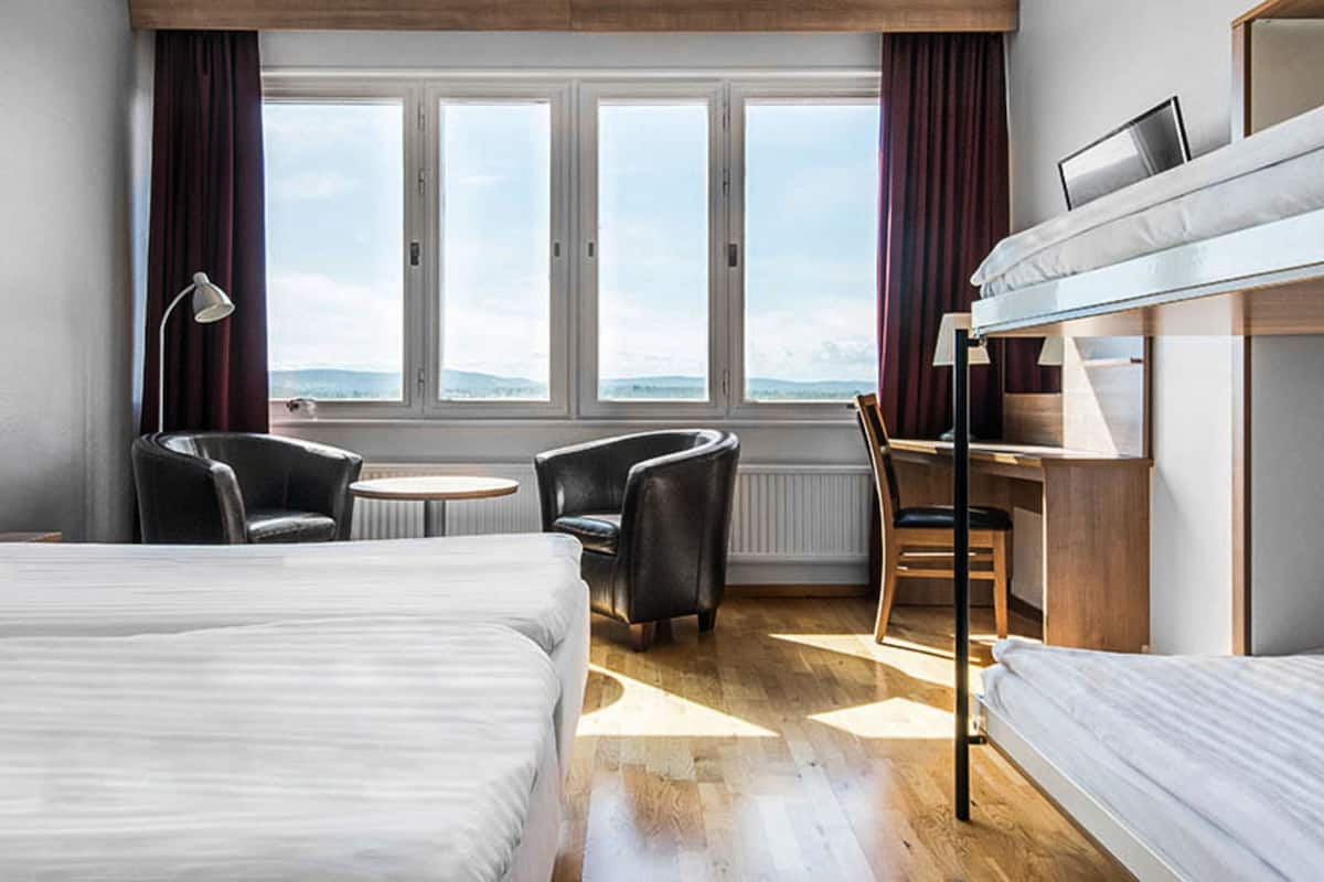 A hotel room with two beds, a desk, a chair, and a view of the mountains through large windows.