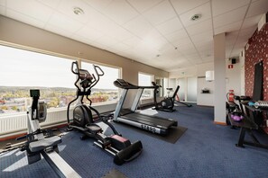 A gym with treadmills, stationary bikes, and a rowing machine, with a view of a cityscape through large windows. - Quality Hotel Bodensia (Boden)