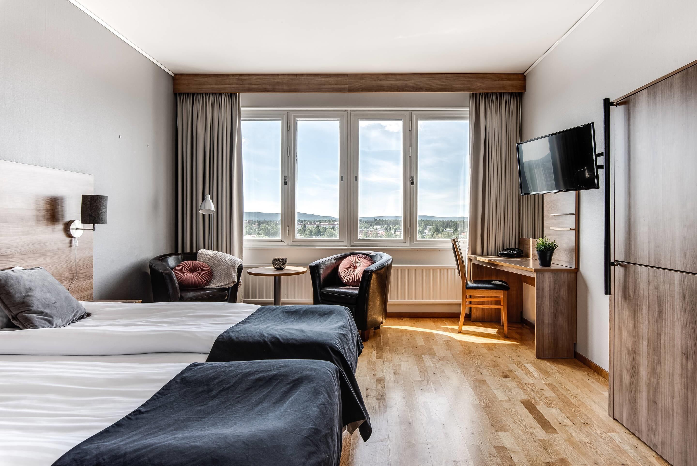 A hotel room with two beds, a desk, a chair, a TV, and a large window with a view.