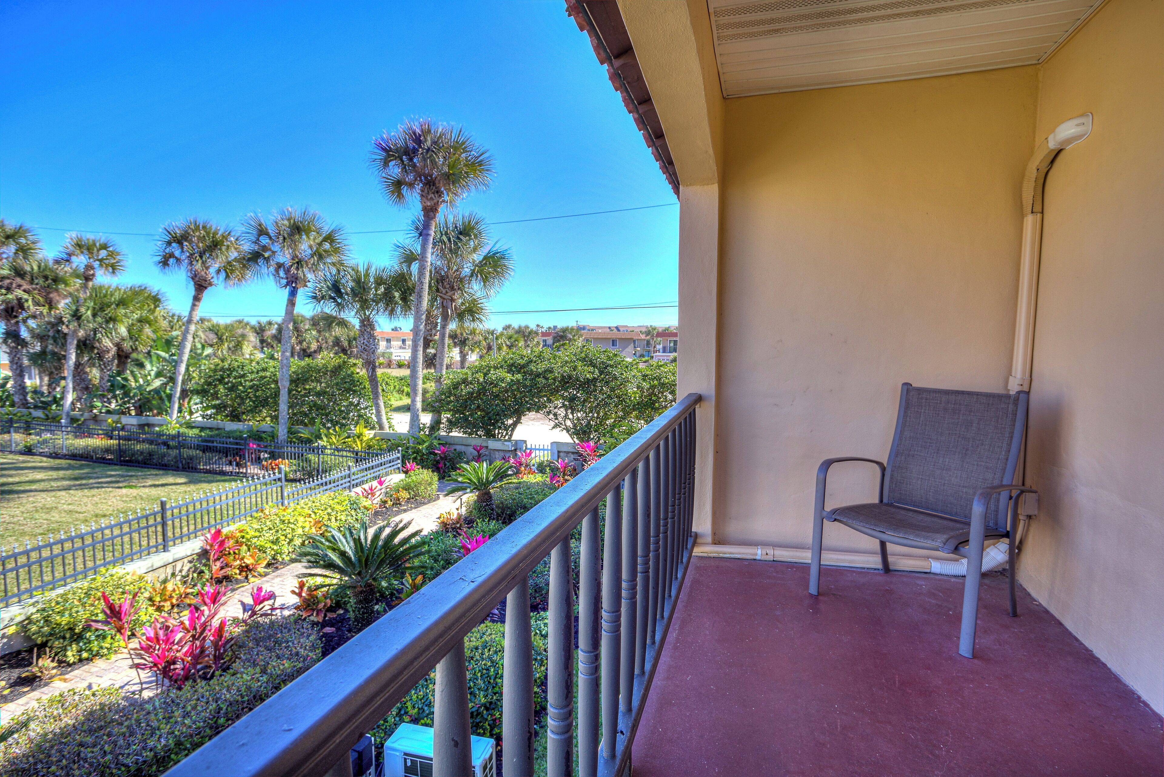 A balcony with a chair, a view of a landscaped garden with palm trees, and a clear blue sky.