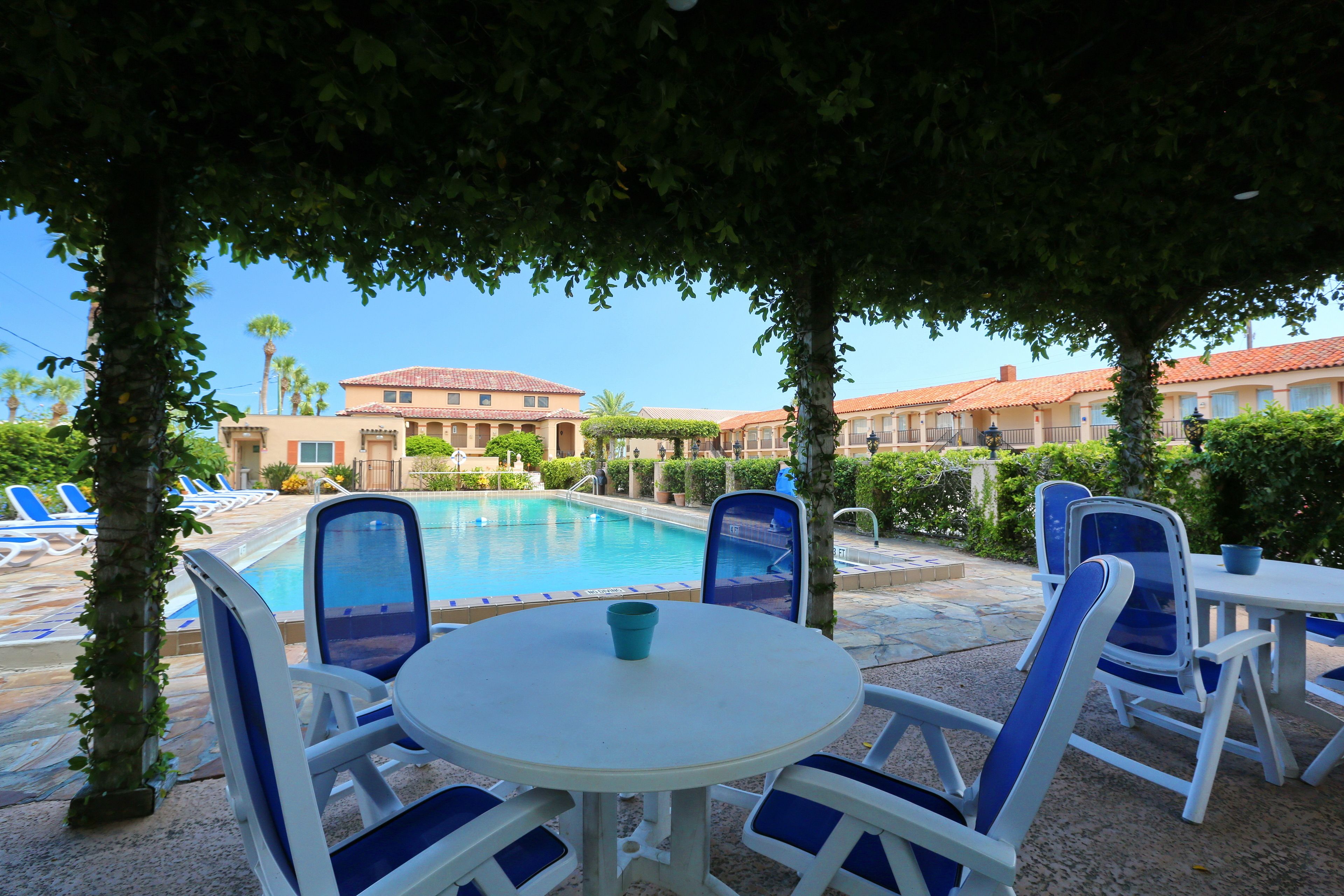 A poolside area with lounge chairs and a table, surrounded by trees and buildings with red-tiled roofs.
