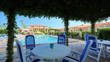 Zona de piscina con sillones de descanso y una mesa, rodeada de árboles y edificios con tejados de tejas rojas.