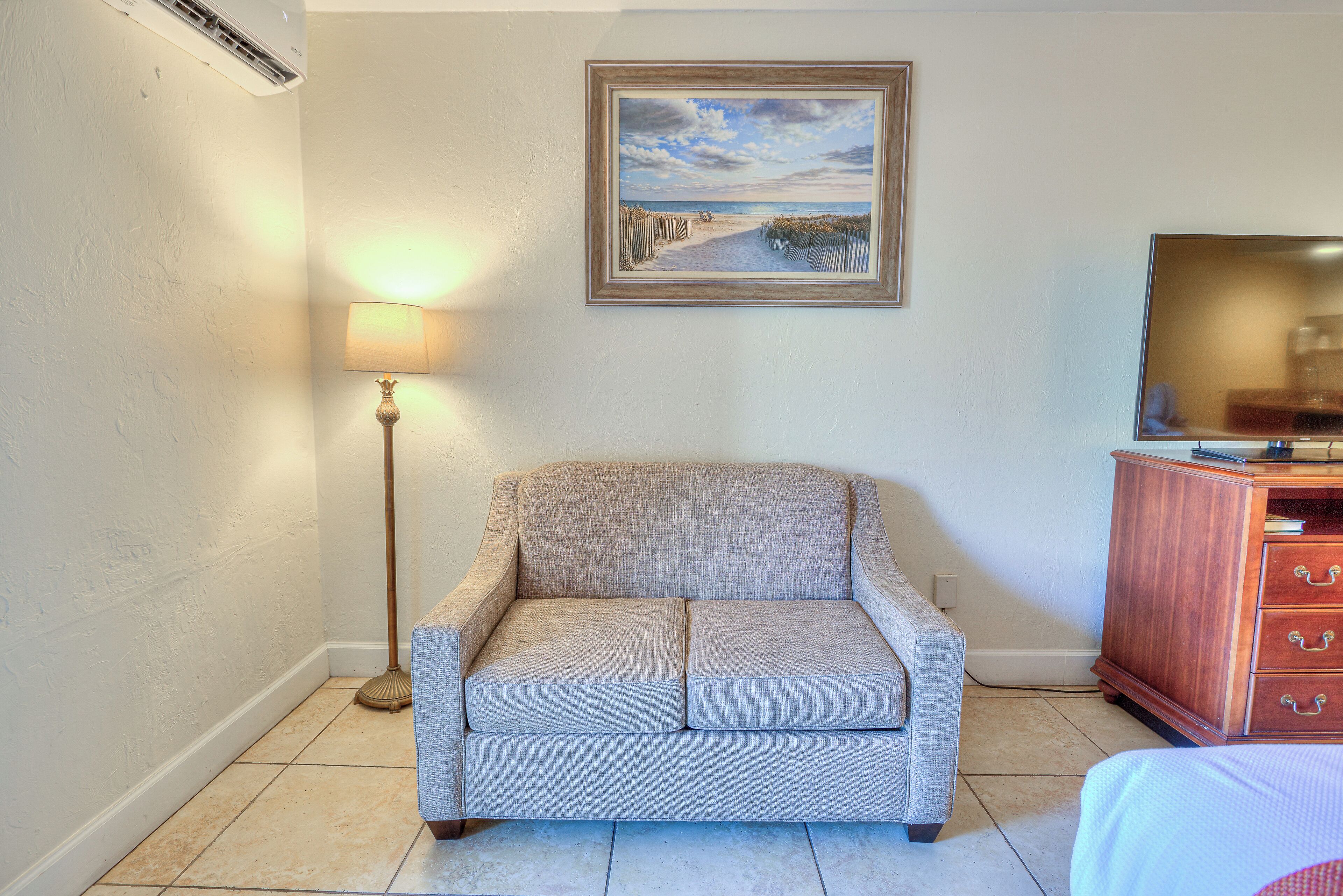 A living room with a beige sofa, a painting of a beach scene, a lamp, and a TV on a wooden cabinet.