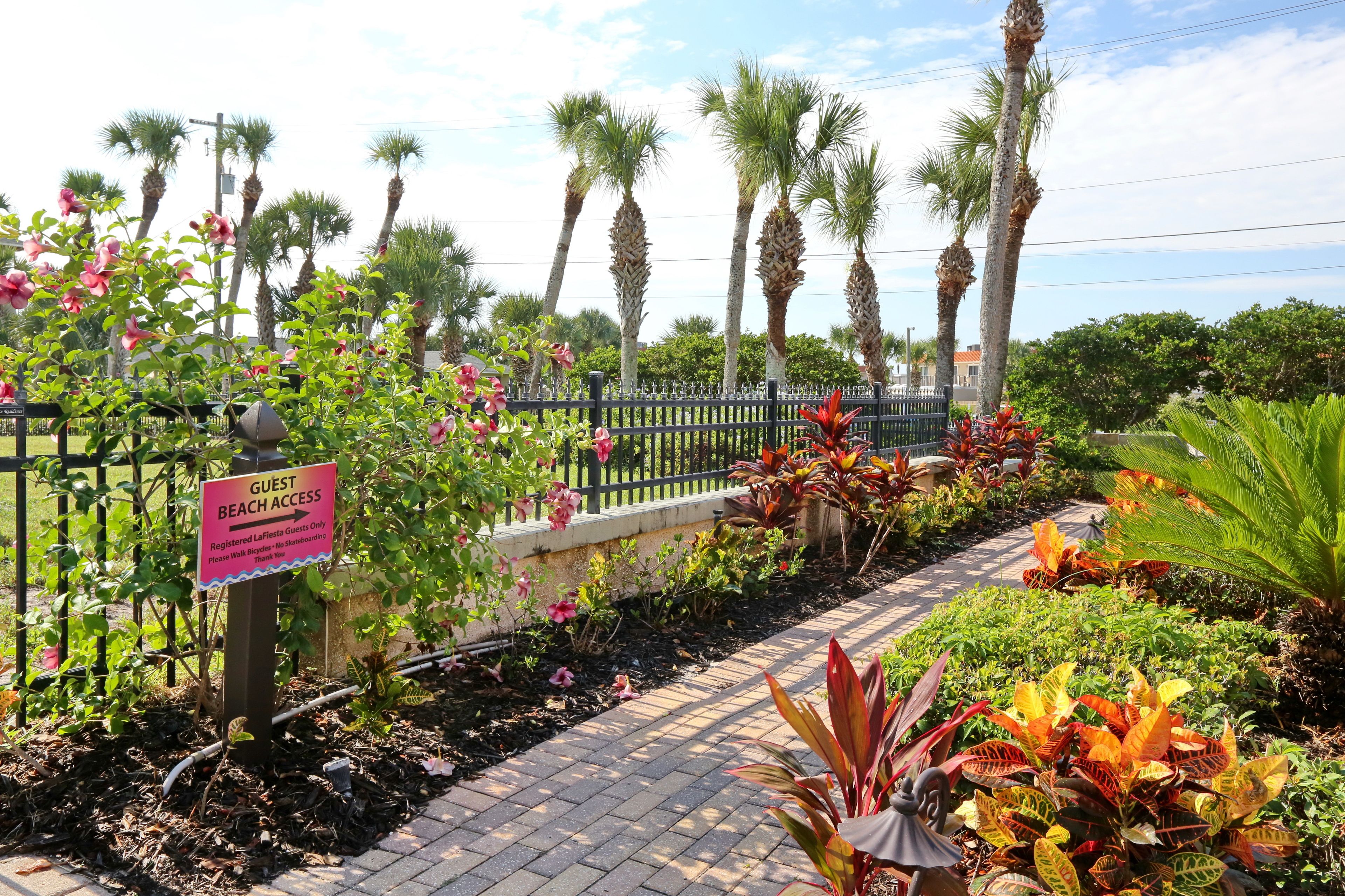 A garden pathway with a sign indicating guest beach access, surrounded by various plants and palm trees.