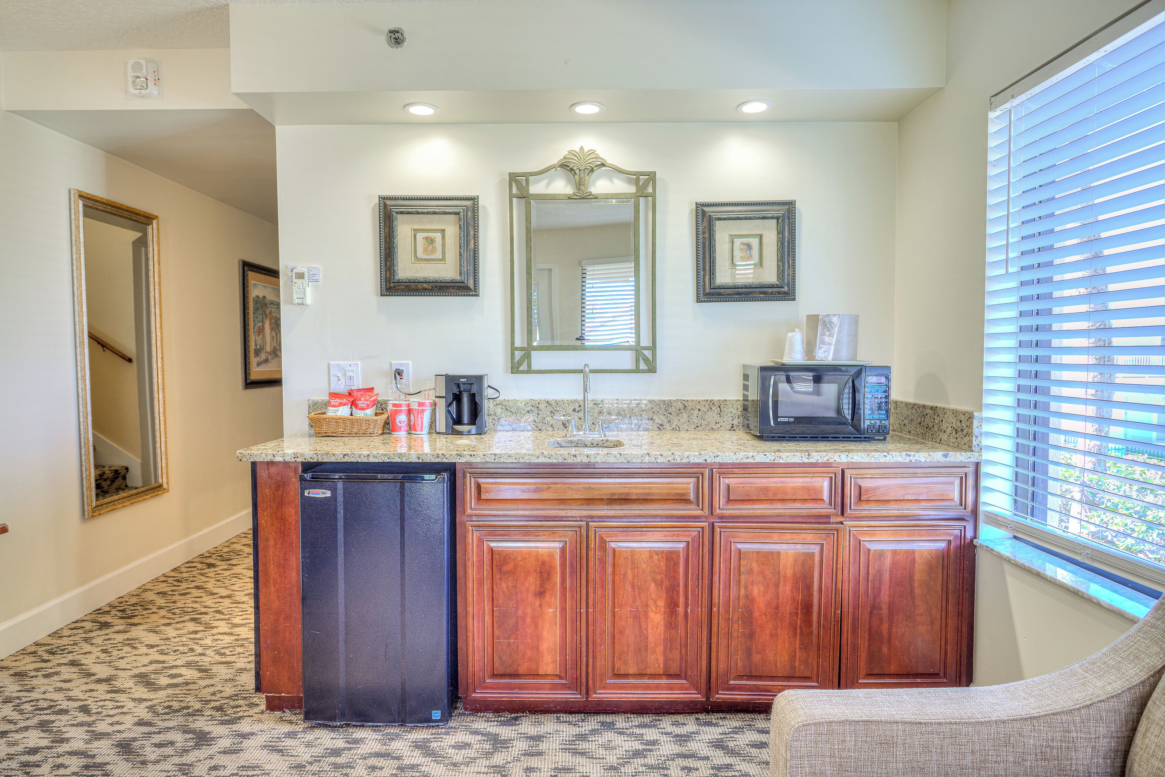 A kitchen with wooden cabinets, a refrigerator, a microwave, and a sink. There are framed pictures on the wall and a window with blinds.
