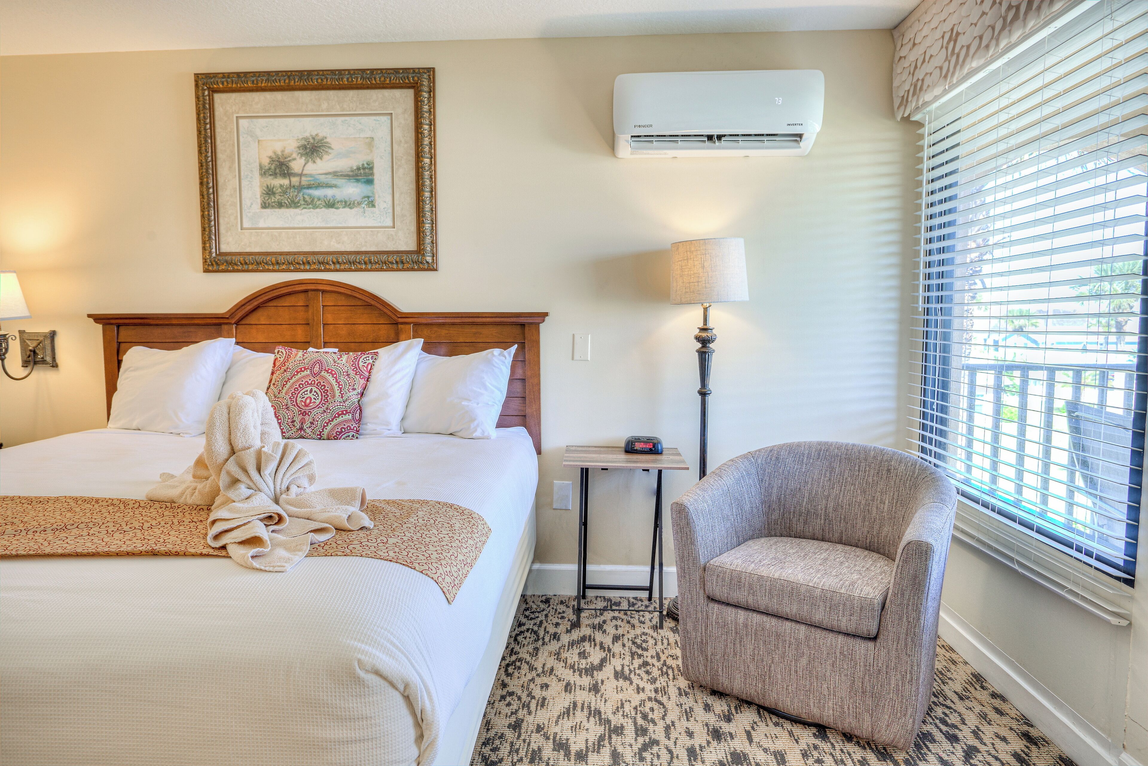 A hotel room with a bed, a chair, a bedside table with a lamp, an air conditioner, and a window with blinds.