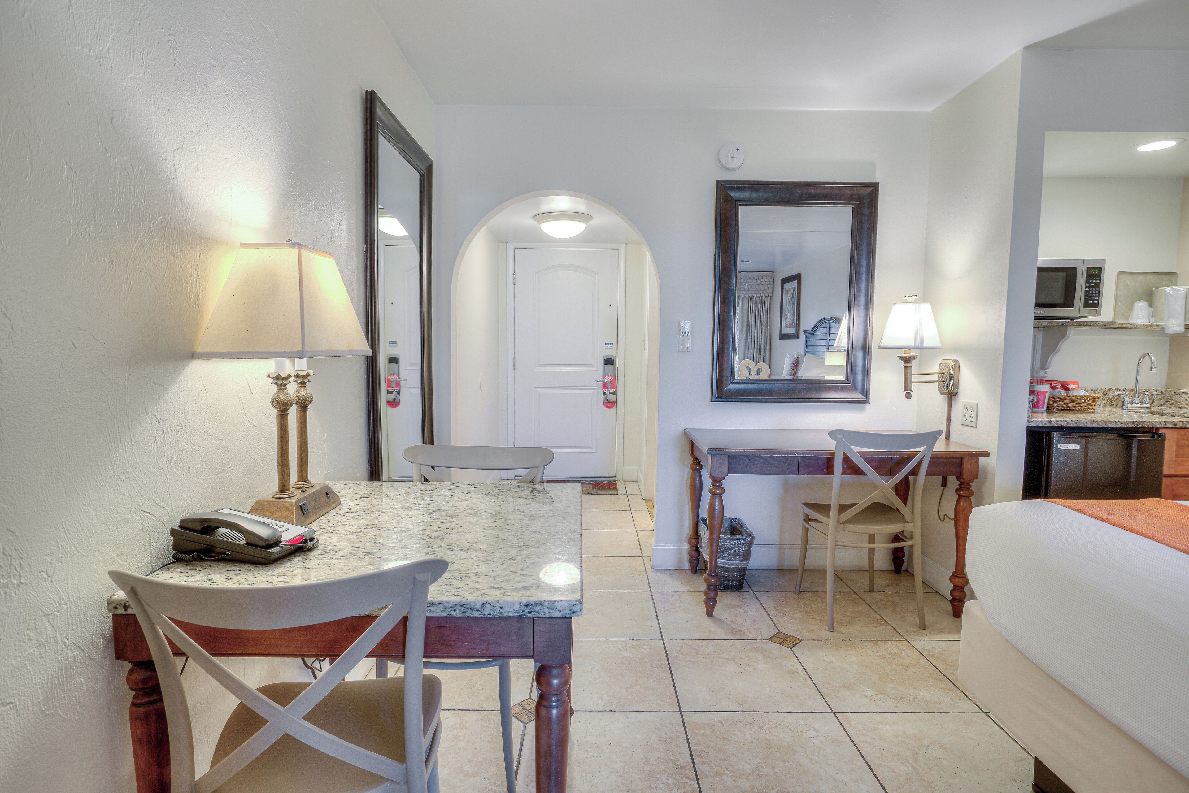 A hotel room with a dining table, chairs, a bed, a desk with a chair, a microwave, and a sink.