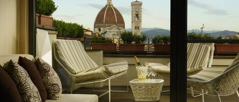 A balcony with a view of a historic building and mountains. There are lounge chairs, a small table with a bottle and glasses, and a cushion with a floral pattern.
