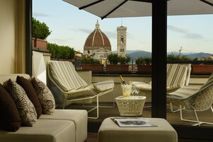A balcony with a view of a historic building and mountains. There are lounge chairs, a small table with a bottle and glasses, and a cushion with a floral pattern.