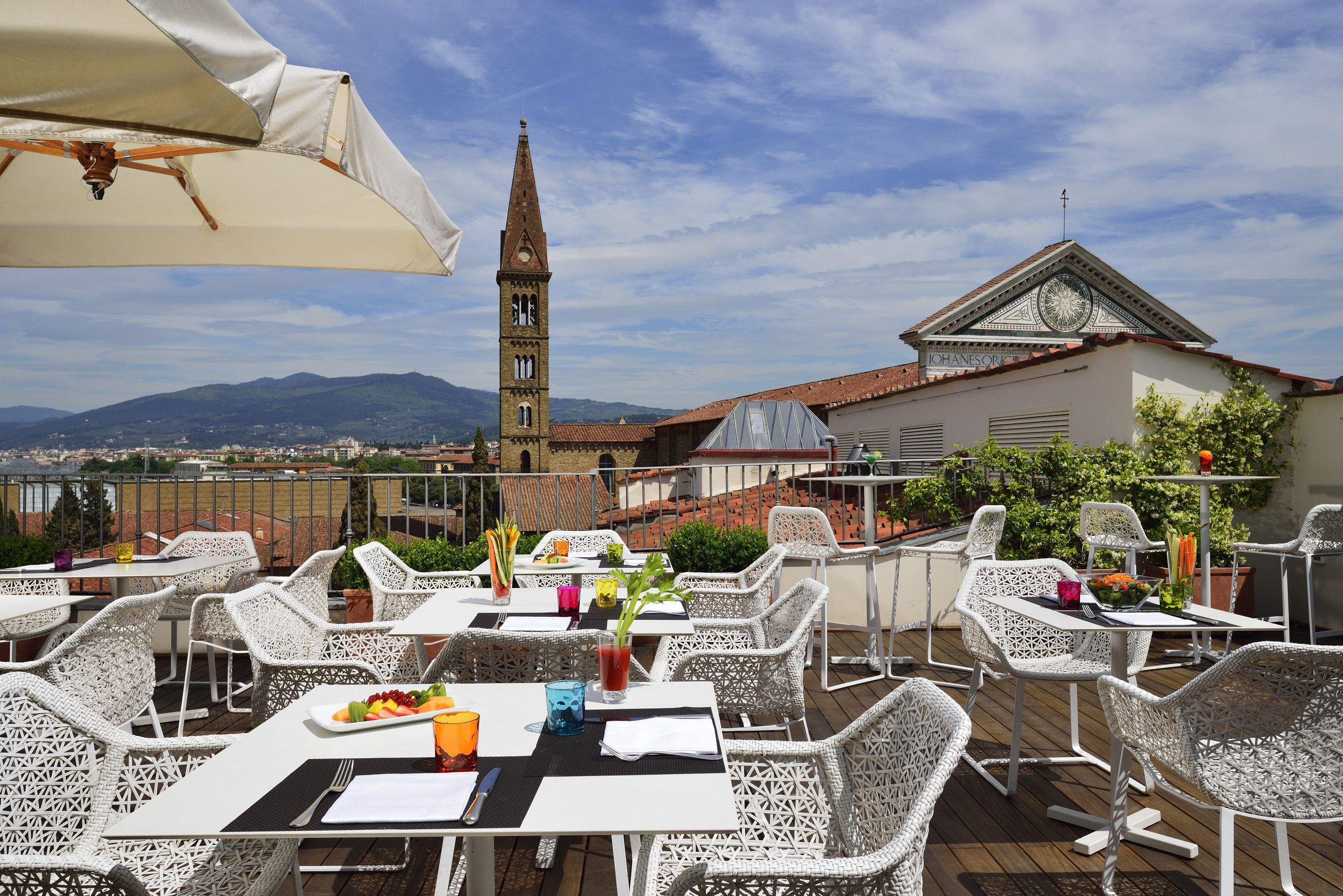 Un espacio para comer al aire libre con muebles de mimbre blanco, vista a una torre de iglesia y un edificio con una torre del reloj.