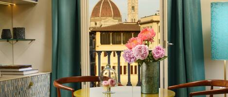 A room with a view of a historic building, a round table with chairs, a vase of flowers, and shelves with decorative items.