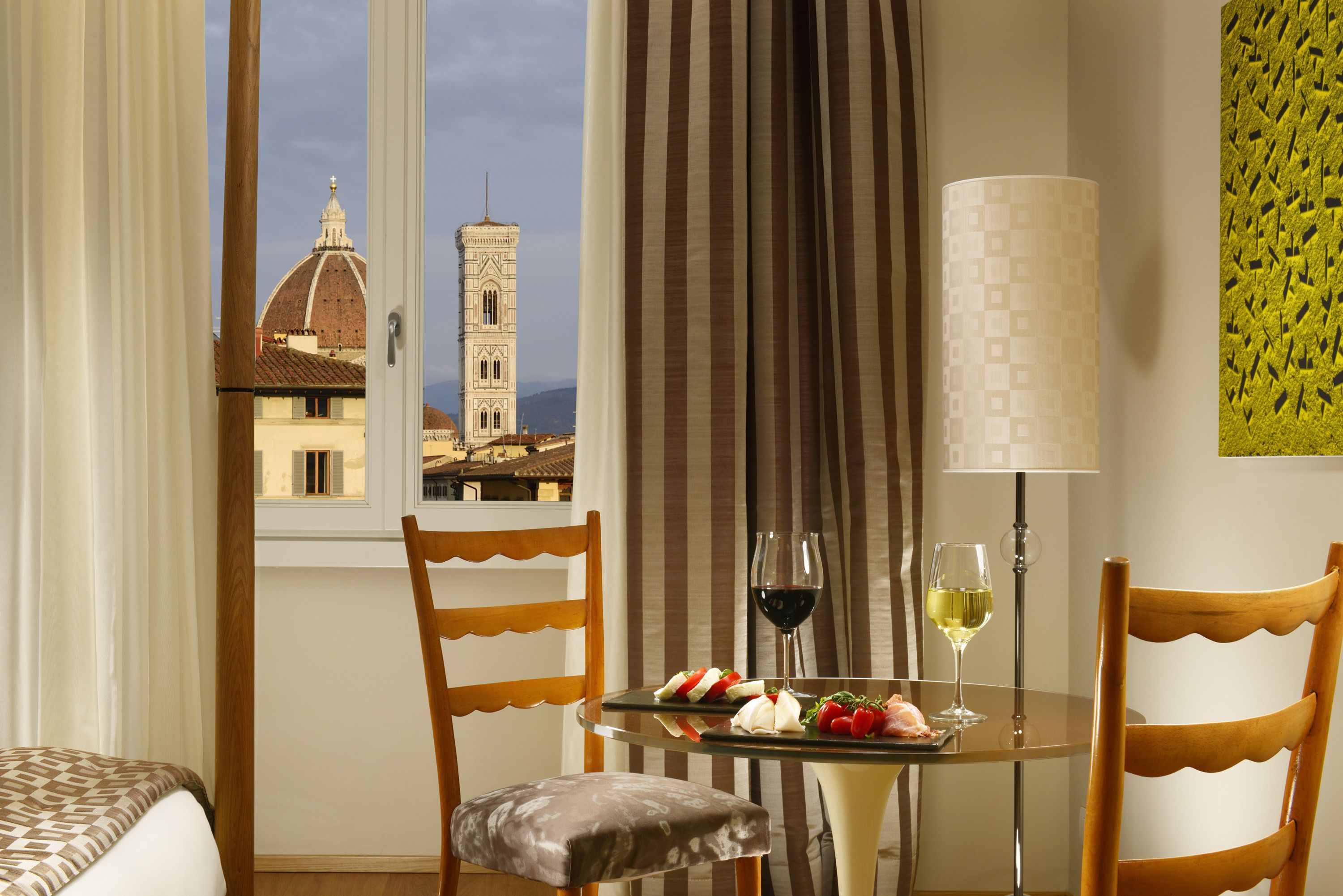A hotel room with a dining table set for two, featuring a view of a historic building and a lamp.