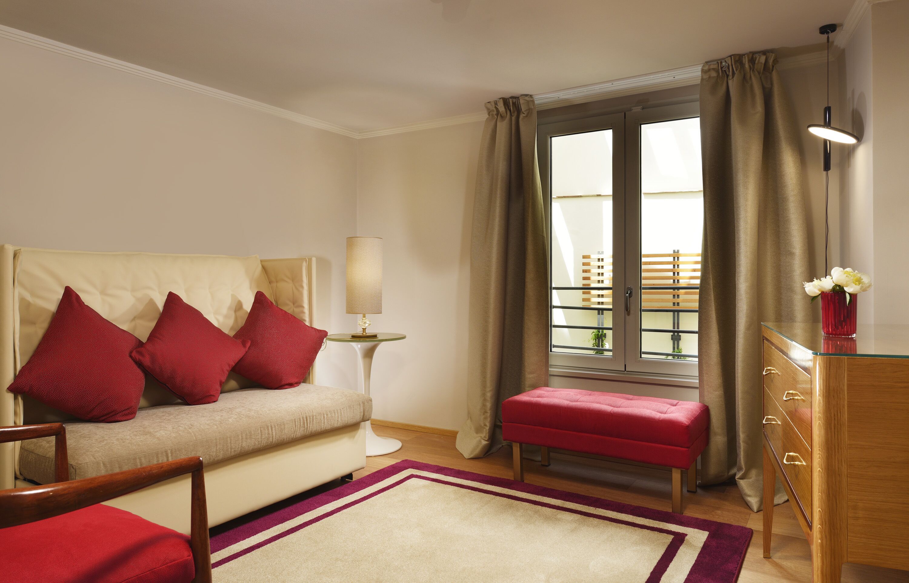 Une chambre d’hôtel avec un canapé, des coussins rouges, une petite table avec une lampe, une commode en bois et une fenêtre avec des rideaux.