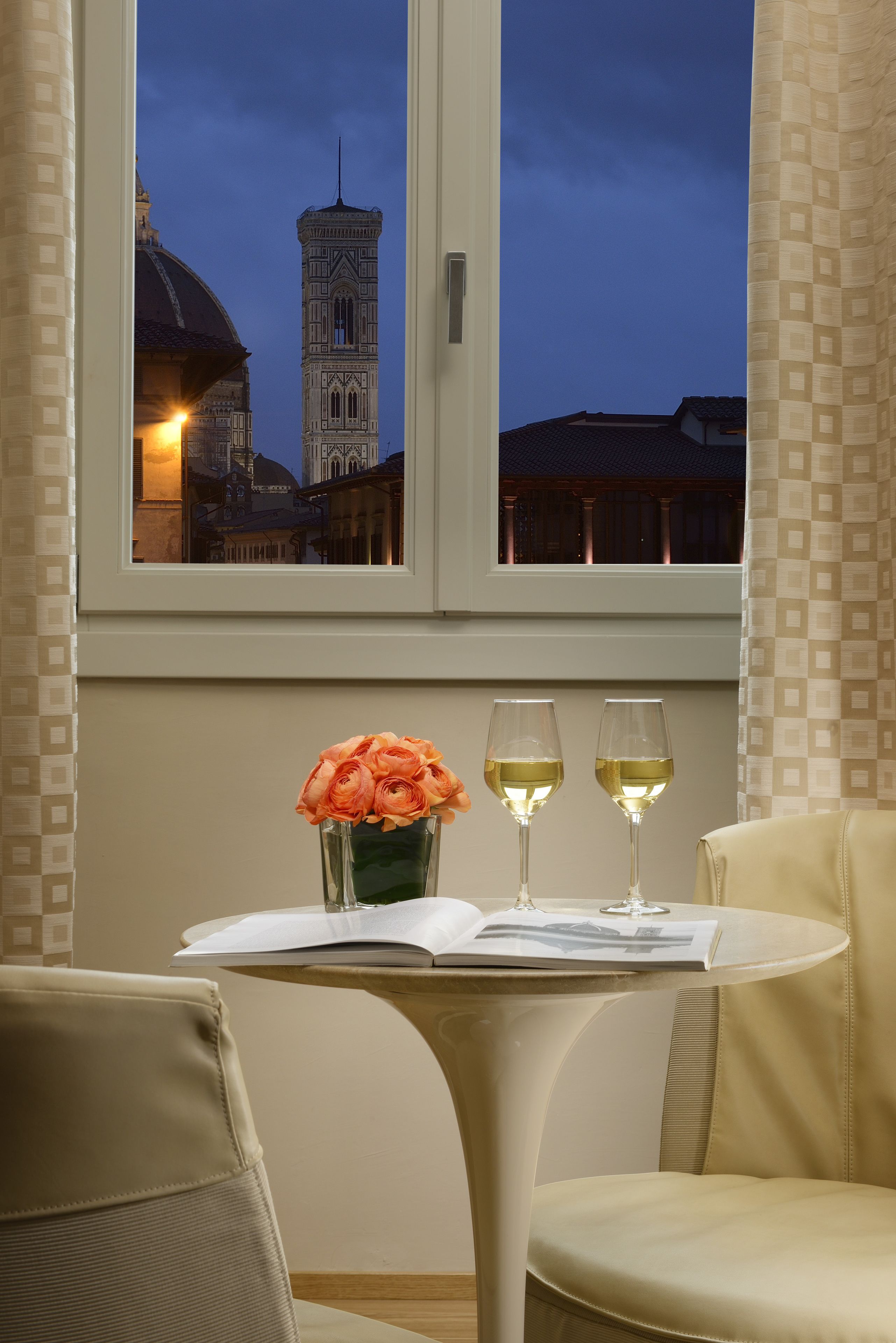 Una habitación con vistas a una torre, una mesa con un jarrón de flores y dos copas de vino, y una silla.