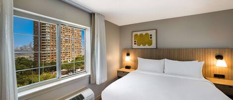 A hotel room with a large bed, two bedside tables with lamps, a view of tall buildings through a window, and a wall-mounted artwork.