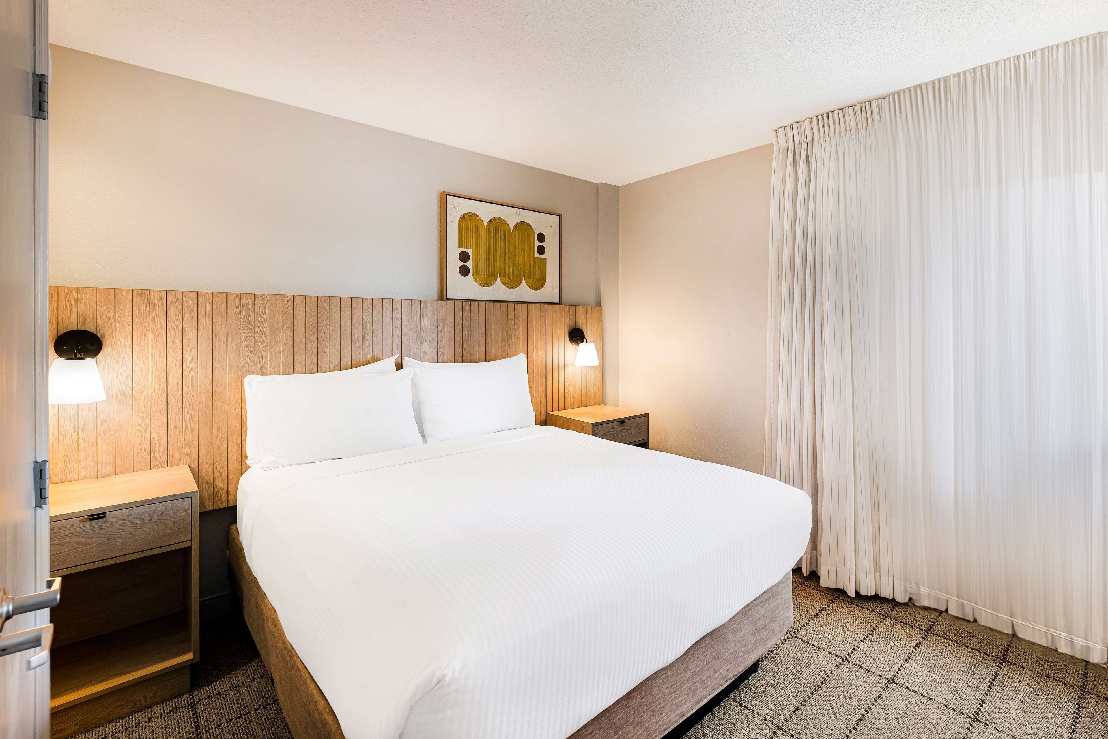 A hotel room with a wooden headboard, a large bed with white linens, two bedside tables with lamps, and a large window with sheer curtains.