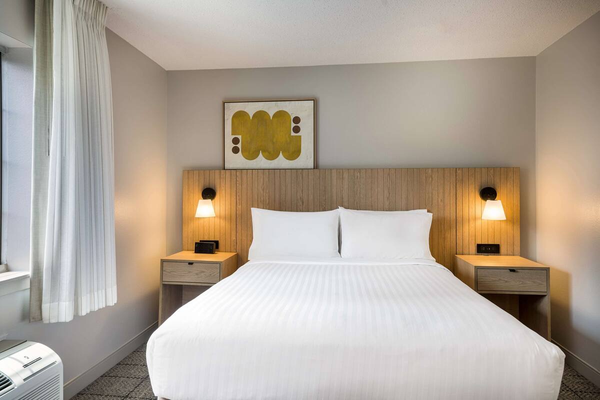 Une chambre d’hôtel avec un grand lit, deux tables de chevet avec des lampes, une œuvre d’art accrochée au mur et une fenêtre avec des rideaux.