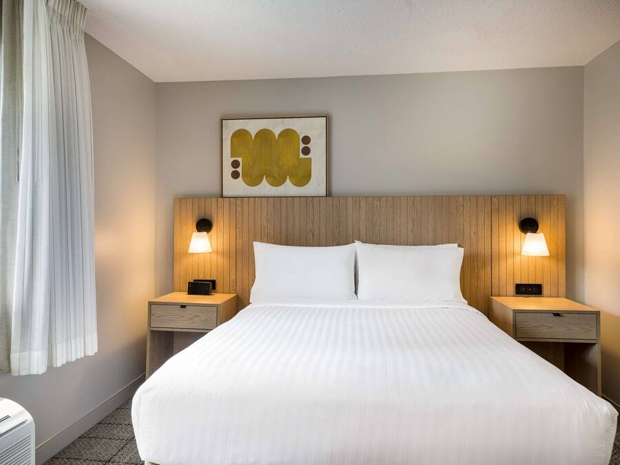A hotel room with a large bed, two bedside tables with lamps, a wall-mounted artwork, and a window with curtains.