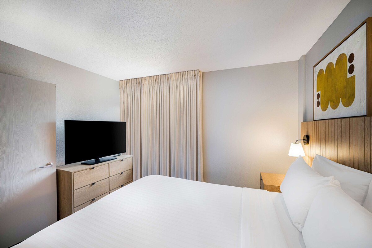 A hotel room with a bed, a TV on a wooden dresser, a lamp, and a large artwork on the wall.
