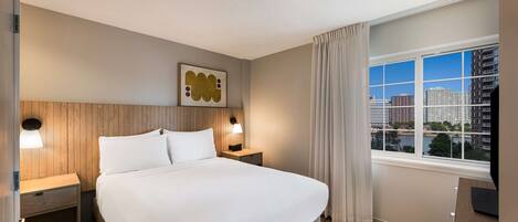 Habitación de hotel con una cama grande, cabecero de madera, dos mesitas de noche con lámparas, vista a la ciudad a través de una ventana y un cuadro en la pared.