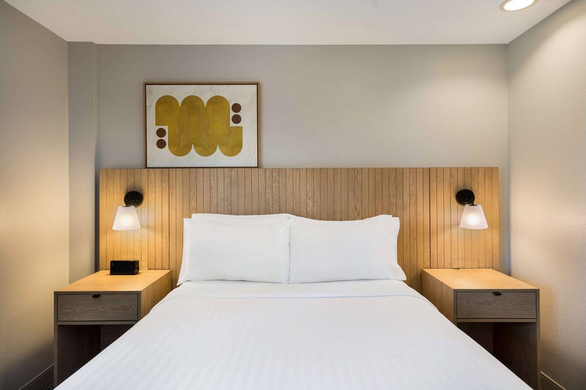 Une chambre d’hôtel avec une tête de lit en bois, deux tables de chevet avec des lampes, un lit avec une literie blanche et une œuvre d’art abstraite encadrée au mur.
