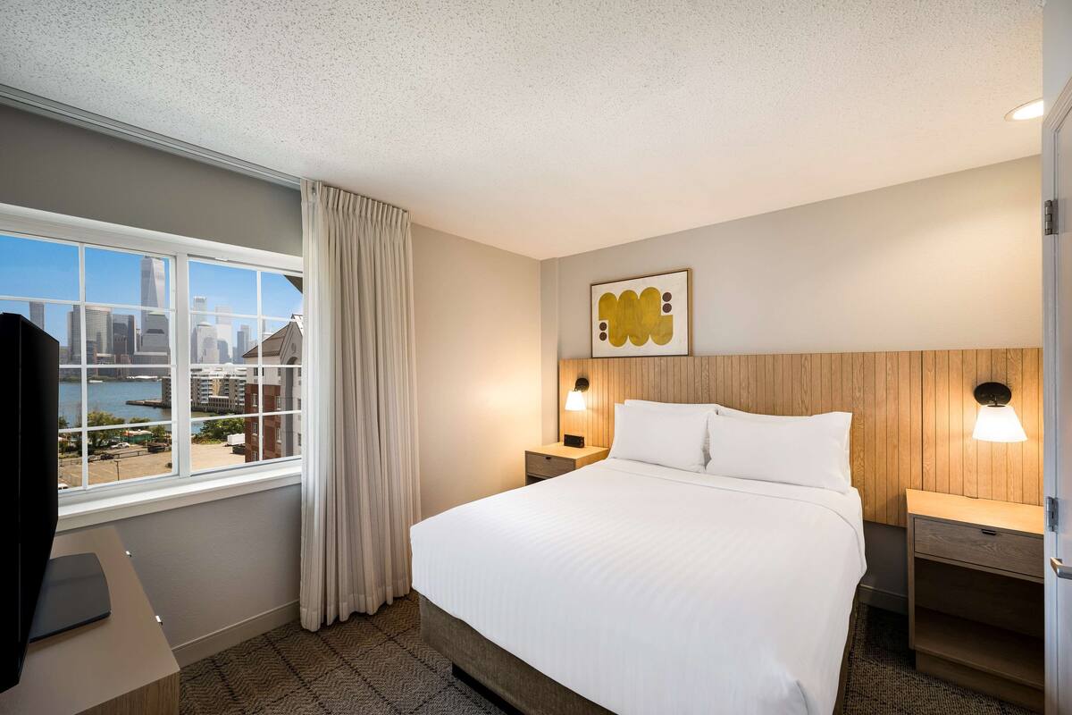 Une chambre d’hôtel avec un grand lit, une table de chevet avec une lampe, une tête de lit en bois, une vue sur le paysage urbain par la fenêtre et un tableau encadré au mur.