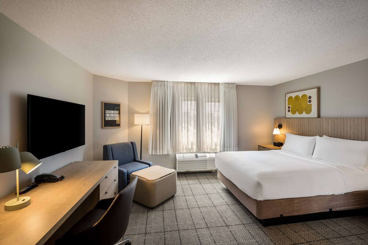 Une chambre d’hôtel avec un lit, un bureau doté d’une lampe, une chaise, un téléviseur et une fenêtre avec des rideaux.