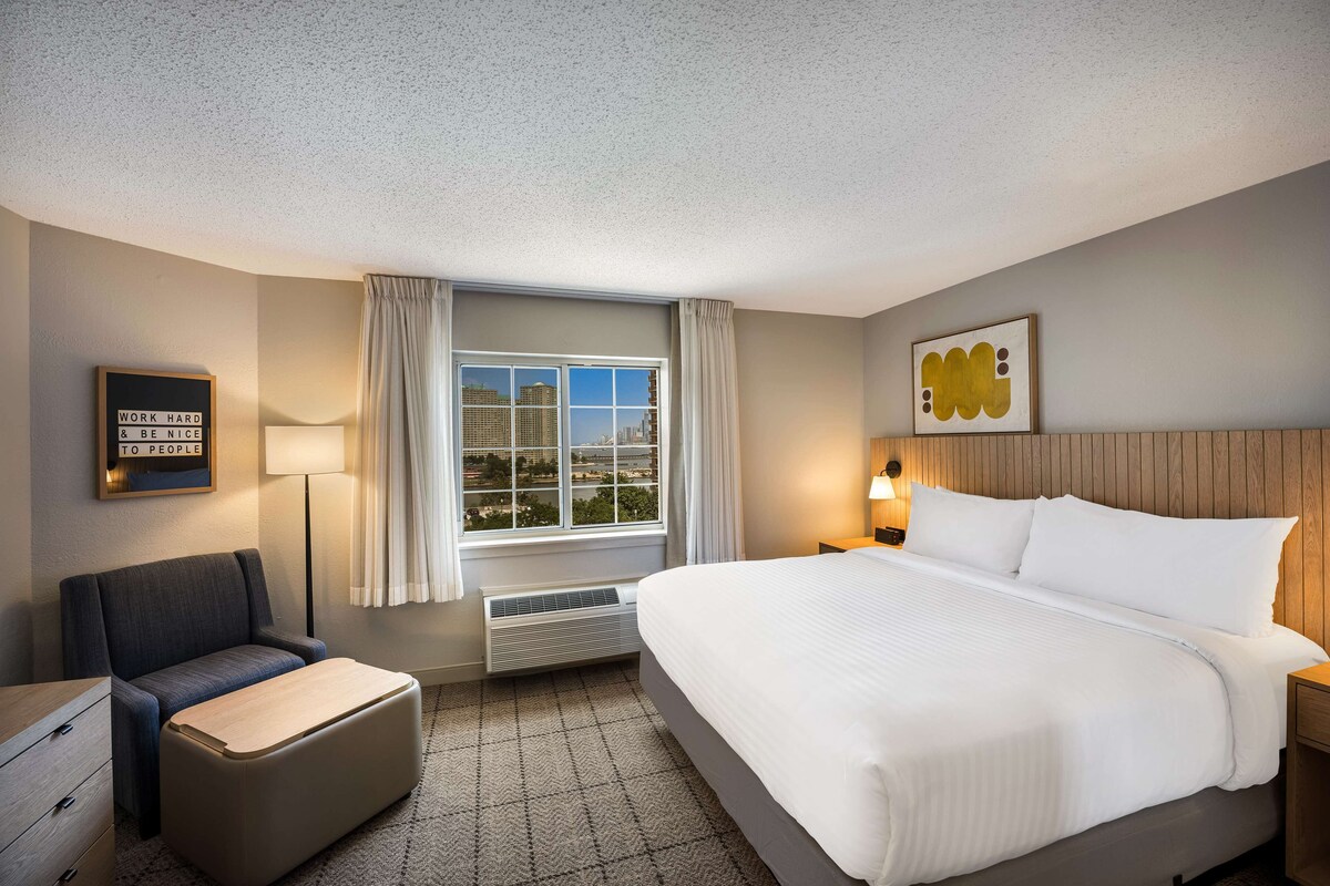 A hotel room with a large bed, a chair, a desk, a lamp, and a view of the city through the window.