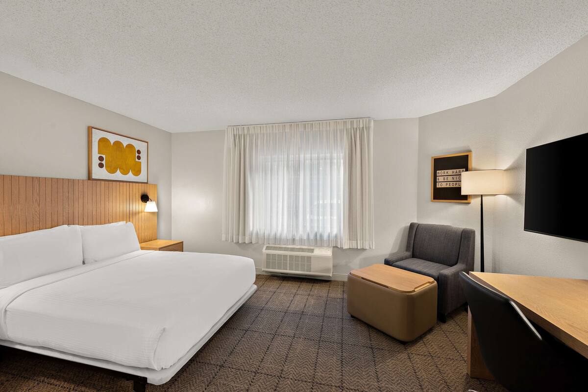 Une chambre d’hôtel comprenant un grand lit, un bureau, une chaise, un téléviseur à écran plat, une lampe fixée au mur, une fenêtre avec des rideaux et un cadre contenant une citation inspirante.
