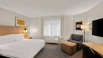 A hotel room with a large bed, a desk, a chair, a flat-screen TV, a wall-mounted lamp, a window with curtains, and a framed motivational quote.