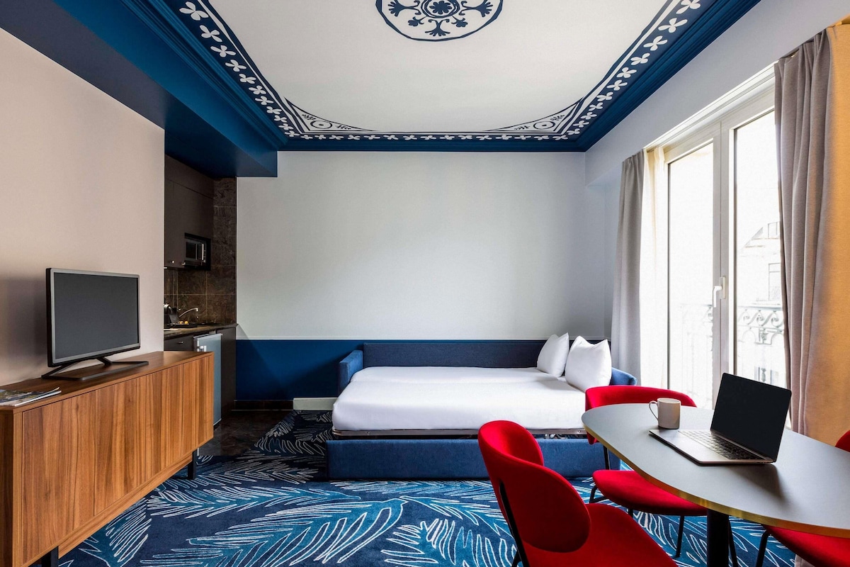 Una habitación de hotel con cama, escritorio, computadora portátil, televisión y ventana con cortinas.