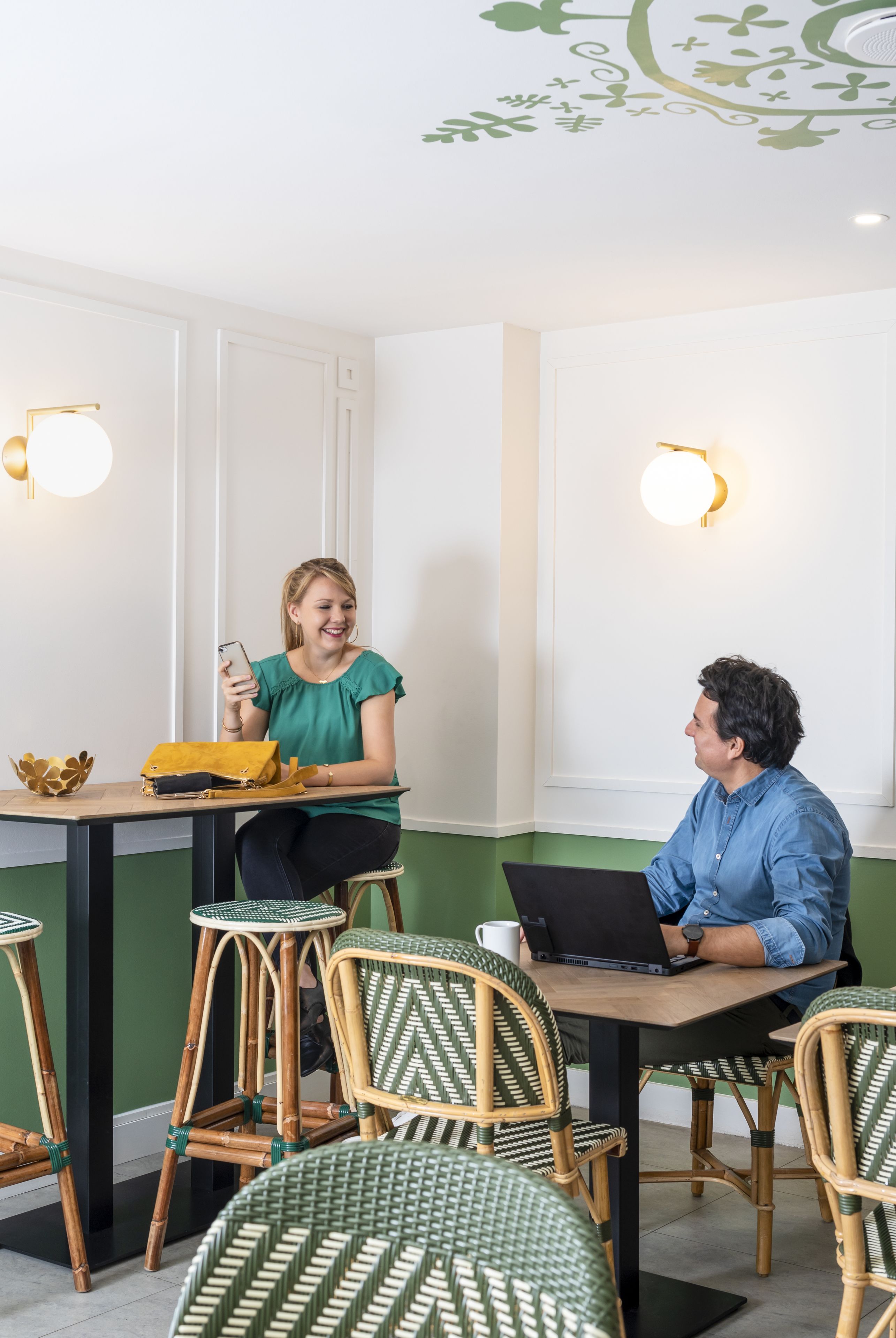 Ein Zimmer mit grüner Wand, weißer Decke und zwei Wandleuchten. Ein Tisch mit einem Laptop und eine Person, die ein Smartphone benutzt.