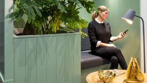A woman sitting on a bench in a room with a green wall, a large plant, and a round table with a lamp and a handbag. - Aparthotel Adagio Paris Haussmann (Paris)