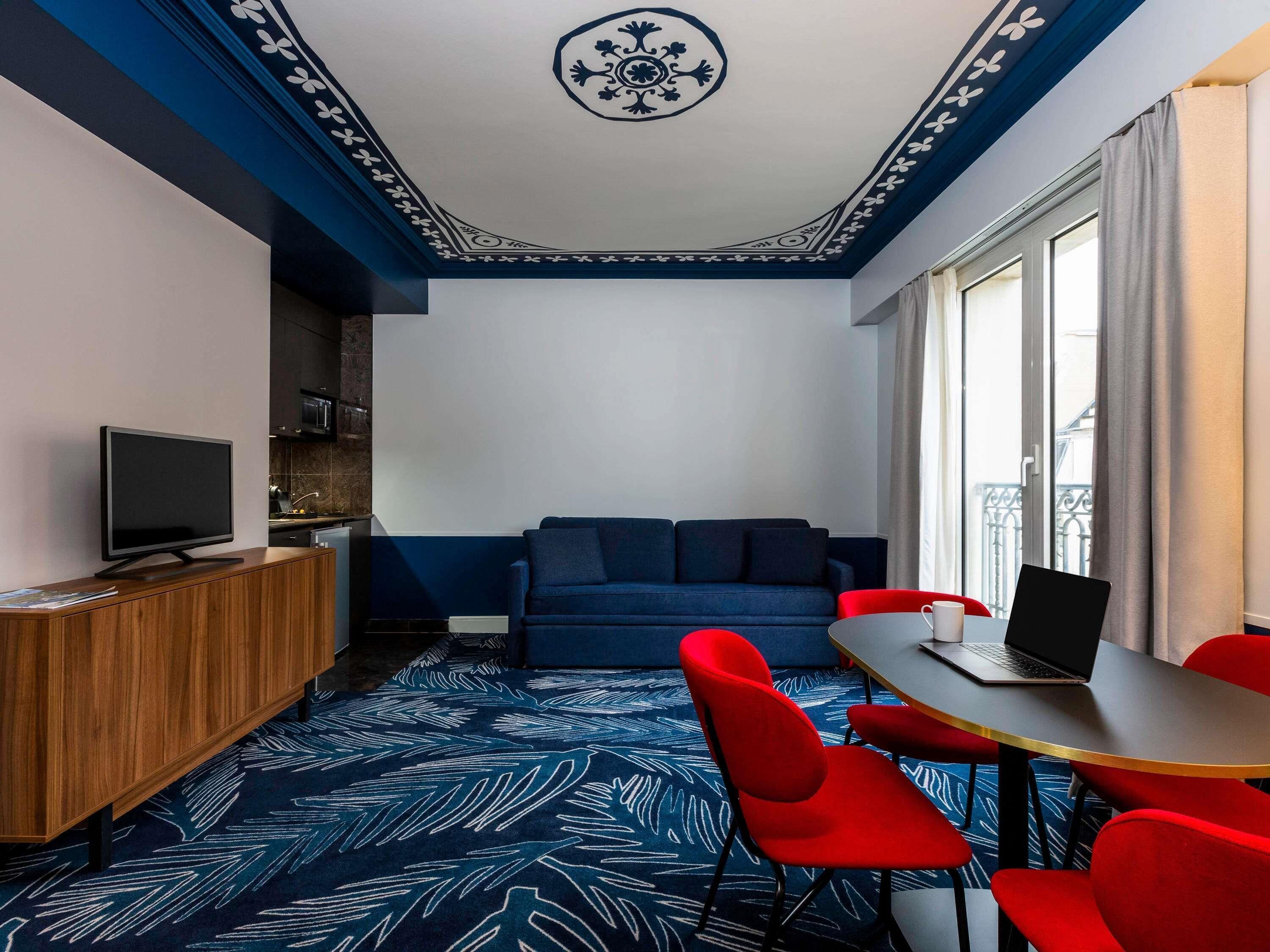 A modern room with a TV, sofa, red chairs, a table with a laptop, and a patterned carpet.